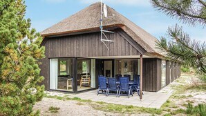 Une maison moderne avec un toit de chaume, de grandes fenêtres en verre et un patio doté de chaises bleues.