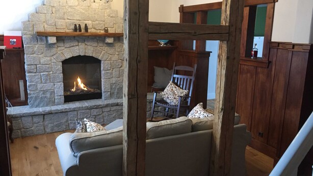 Une cheminée en pierre avec un feu, une chaise en bois et un canapé rembourré dans une pièce aux poutres apparentes et à la balustrade en bois.