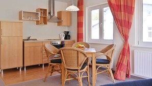Cocina compacta con armarios de madera, una mesa de comedor con sillas de mimbre y una ventana con cortinas rojas.