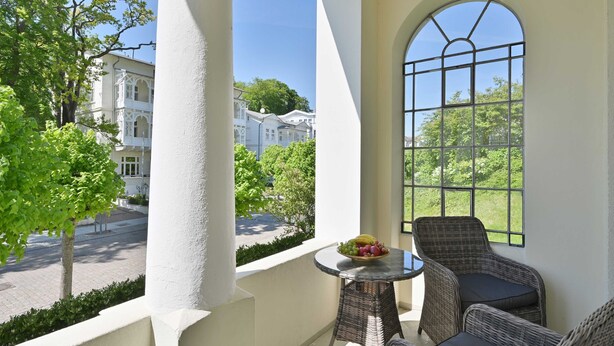 Balcón con mobiliario de mimbre y una mesa con fruta, con vista a una calle con edificios y árboles.