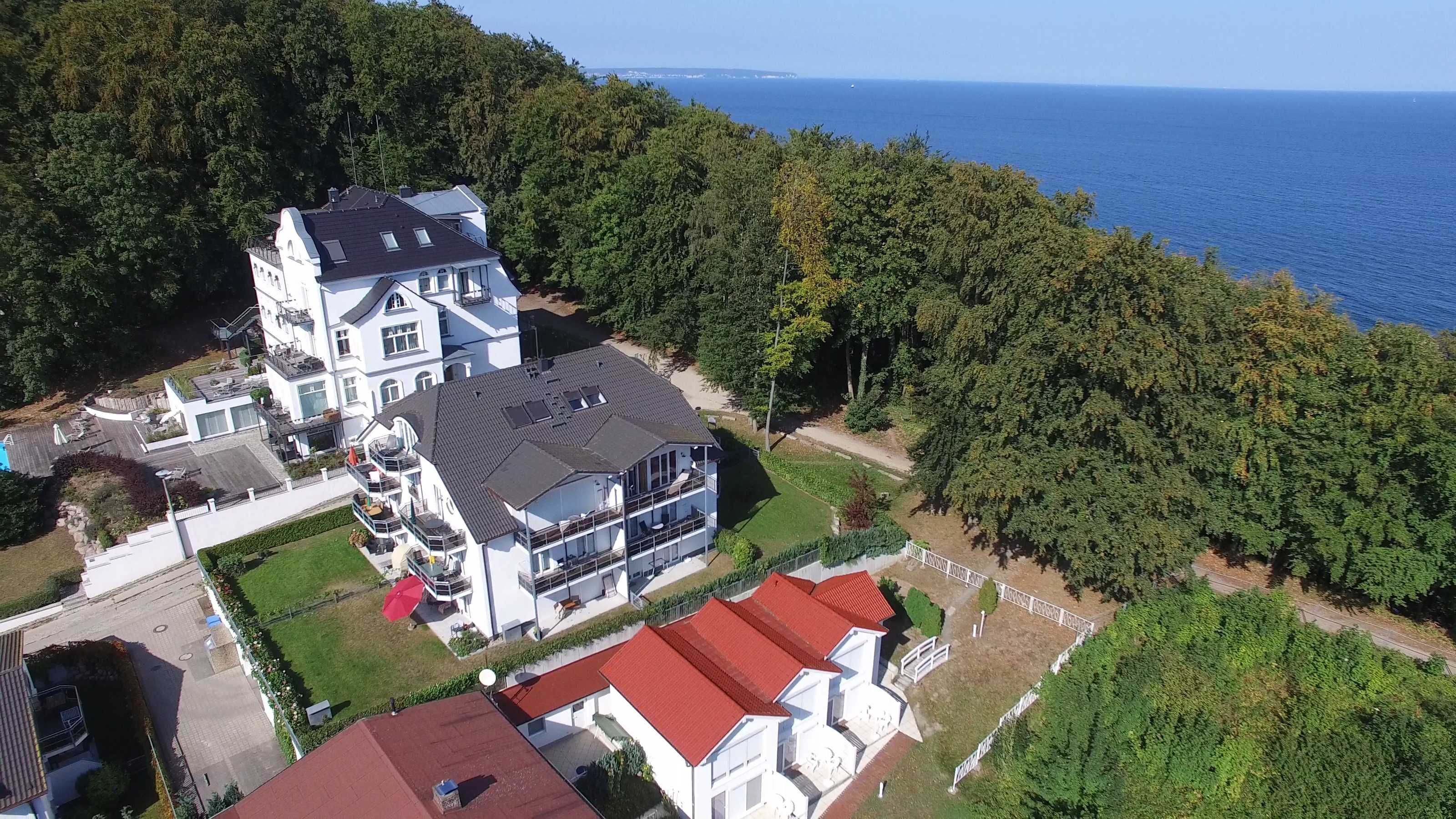A large, multi-story house with a prominent balcony, surrounded by lush greenery and a view of the sea.