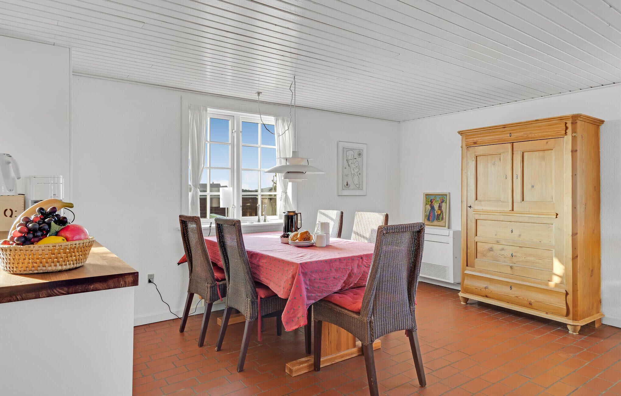 Un coin repas avec une table dressée pour quatre personnes, des chaises en osier, un buffet en bois et un panier de fruits posé sur un comptoir en bois.