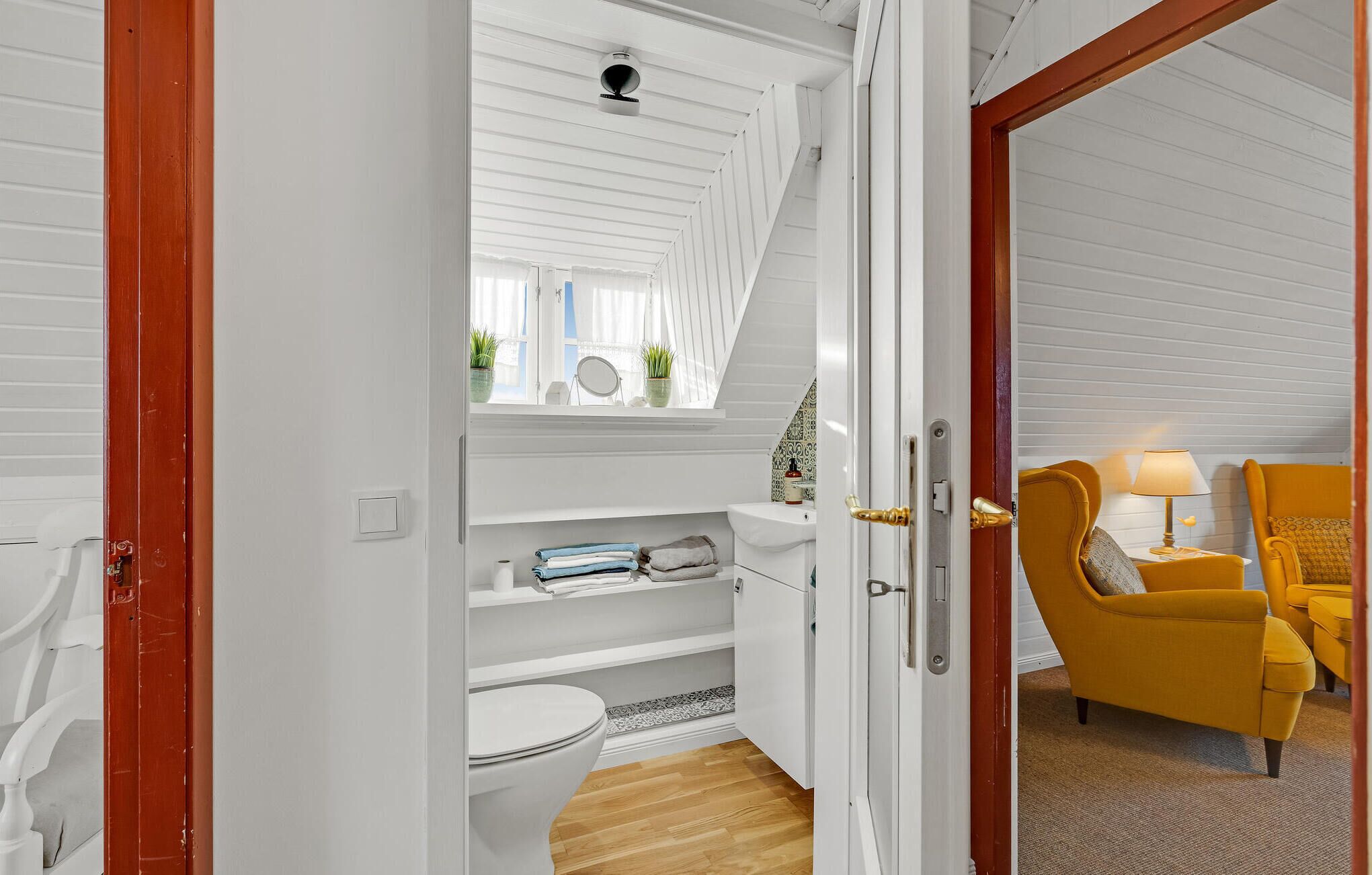 Et værelse med hvide vægge, en trædør, en gul lænestol og et lille bord med en lampe.