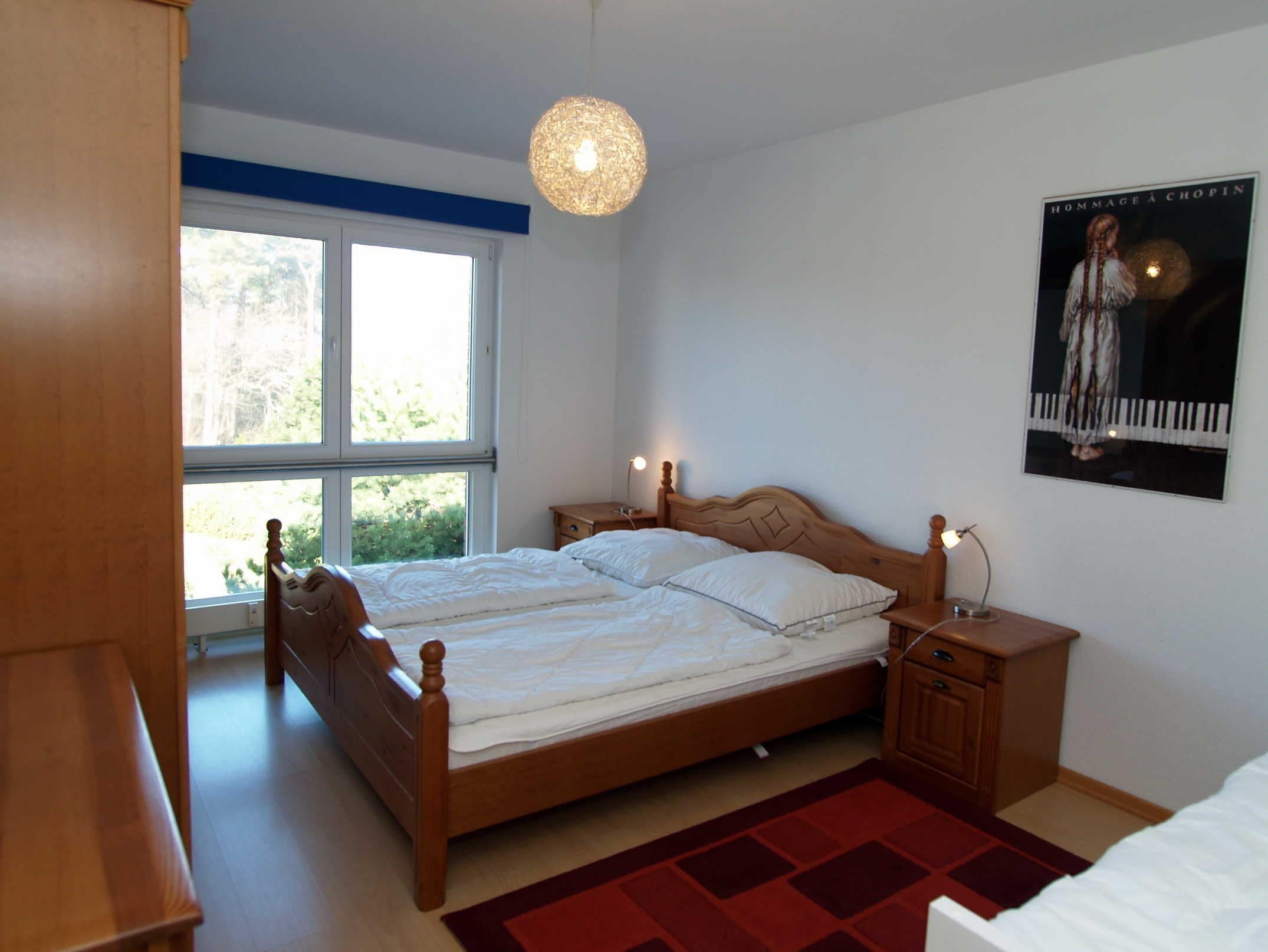 Ein Schlafzimmer mit Doppelbett, Nachttischen, einer Hängeleuchte, einem Fenster und einem Bild an der Wand.