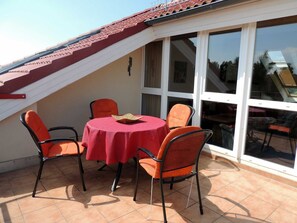 Ein Essbereich mit einem runden Tisch, bedeckt mit einer roten Tischdecke, umgeben von vier orangefarbenen Stühlen. Der Bereich liegt unter einem geneigten Dach mit roten Ziegeln, und im Hintergrund sind große Fenster zu sehen.