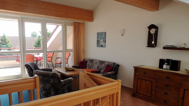 Habitación con una cuna de madera, un sofá, una mesa de comedor y sillas. Se aprecia un ventanal con cortinas, un reloj de pared y un cuadro enmarcado.