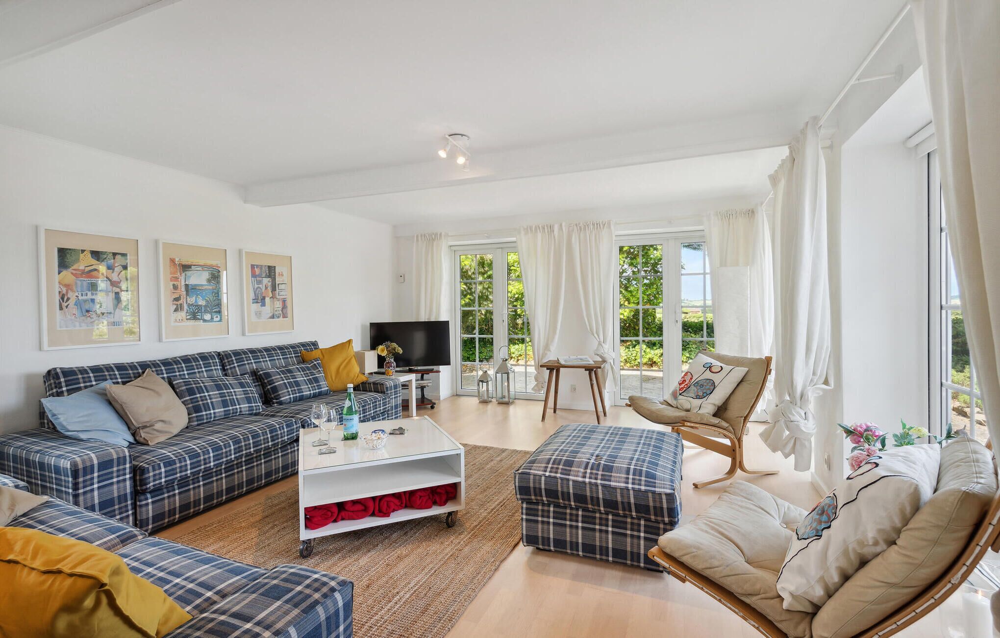 Un salon spacieux comprenant un ensemble de canapés, une table basse et un téléviseur. De grandes fenêtres sont agrémentées de rideaux, un tableau orne le mur et une vue sur la verdure s’offre à l’extérieur.