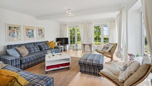 A spacious living room with a sofa set, a coffee table, and a TV. There are large windows with curtains, a painting on the wall, and a view of greenery outside. - 3 bedroom stunning home in Sydals (Sydals)