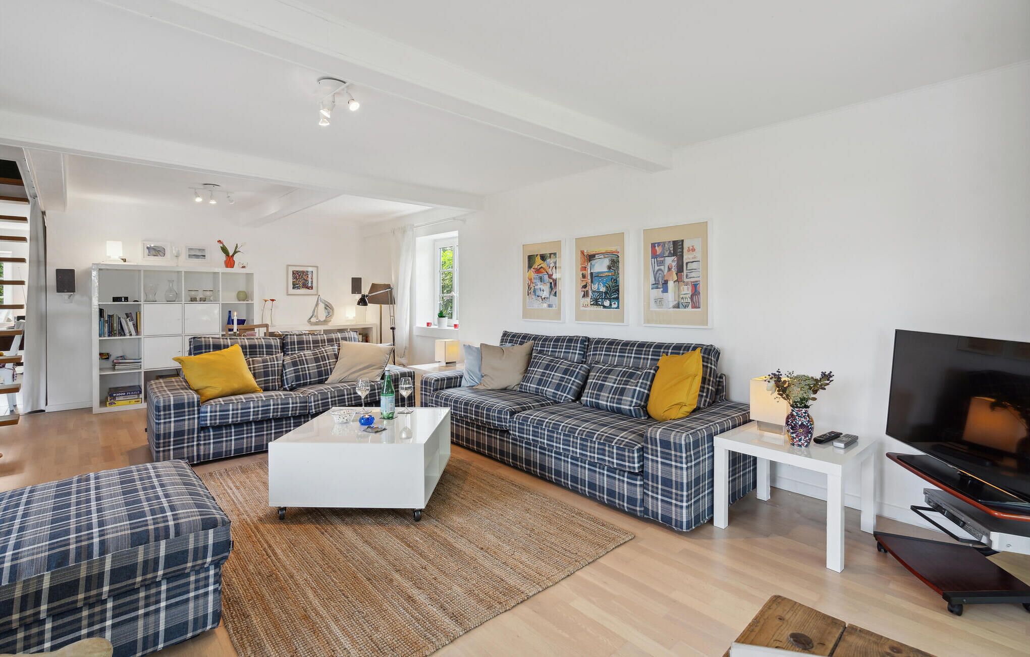 A modern living room with two plaid sofas, a white coffee table, and a flat-screen TV. There is a bookshelf, a lamp, and artwork on the walls.
