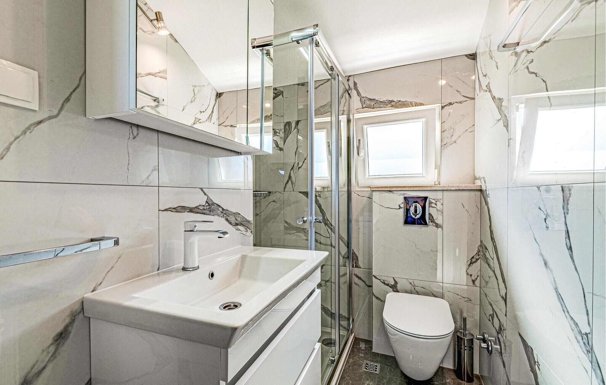 A modern bathroom with a marble backsplash, a glass shower enclosure, a white sink, and a toilet.