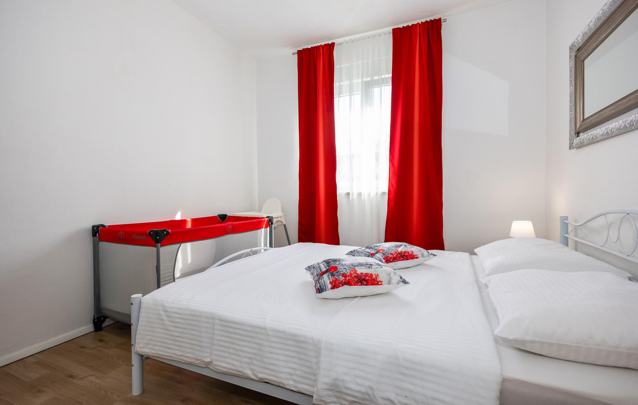A bedroom with a bed, pillows, a red chair, and a window with red curtains.