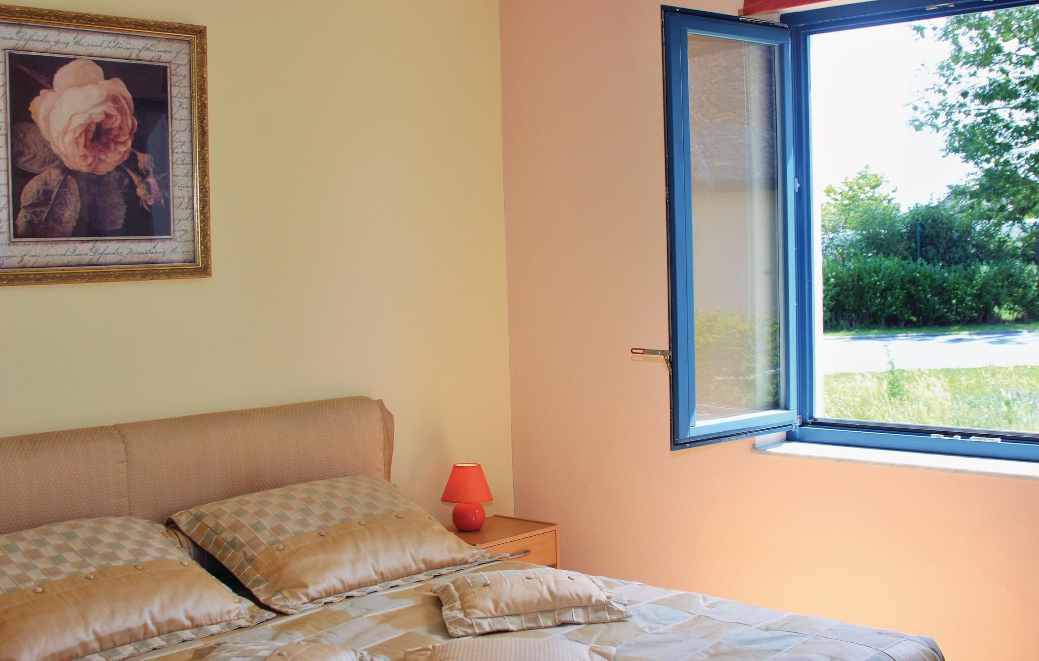 Un dormitorio con cama, almohadas, mesita de noche, lámpara, ventana y un cuadro en la pared.