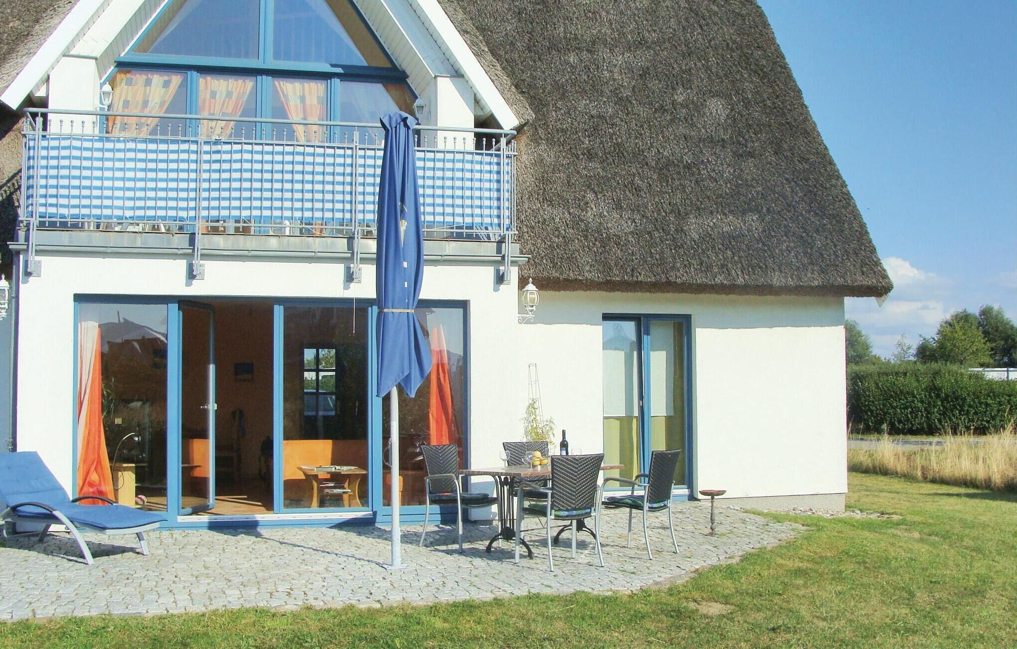 Ein zweigeschossiges Haus mit Strohdach, einem Balkon mit blauer, blau-weiß karierter Markise und einer Terrasse mit Gartenmöbeln.