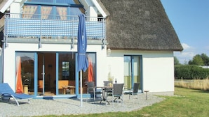 Una casa de dos pisos con techo de paja, un balcón con toldo azul a cuadros y un patio con mobiliario exterior.