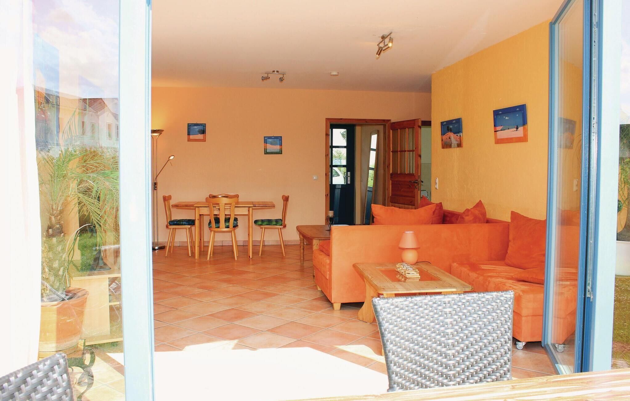 A living room with a sofa, a dining table with chairs, and a small table with a lamp. There are framed pictures on the walls and a door leading outside.
