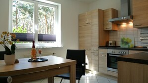 A modern kitchen with wooden cabinets, a dining table with chairs, a window with blinds, and a view of trees outside. - Plötz Vacation Rental - with a Sun Terrace and Electric Car Charging Station - Vacation Rental with (Loddin)