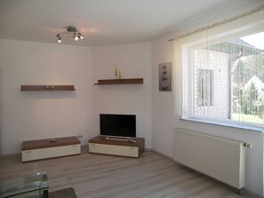 A modern living room with a flat-screen TV, wooden shelves, a glass coffee table, and a large window with blinds.