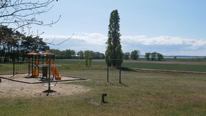 A playground with a swing set and climbing structures, surrounded by open fields and a tall tree. - Plötz Vacation Rental - with a Sun Terrace and Electric Car Charging Station - Vacation Rental with (Loddin)