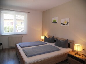 A bedroom with two beds, a window with blinds, a radiator, and two wall-mounted lamps.