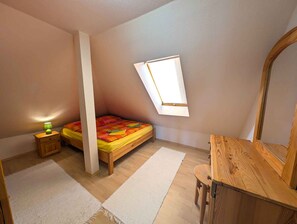 A bedroom with a slanted ceiling, a bed with a colorful bedspread, a bedside table with a lamp, a wooden dresser, and a large mirror.