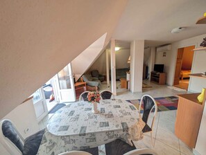 A dining area with a table set for two, a flower vase, and a view of a living room with a sofa and a television. - Holiday apartment with garden, balcony and air conditioning (Balatonfenyves)