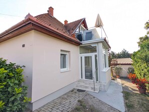 A single-story house with a sloped roof, a balcony with a glass enclosure, and a small front porch. - Holiday apartment with garden, balcony and air conditioning (Balatonfenyves)