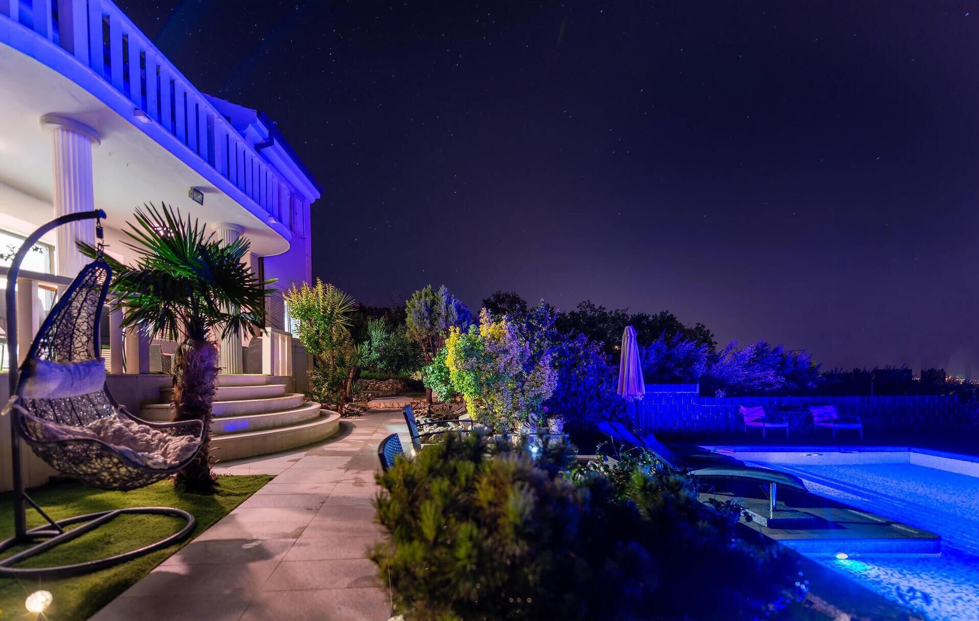 Una vista nocturna de una propiedad lujosa con piscina, asientos exteriores y una escalera bien iluminada.