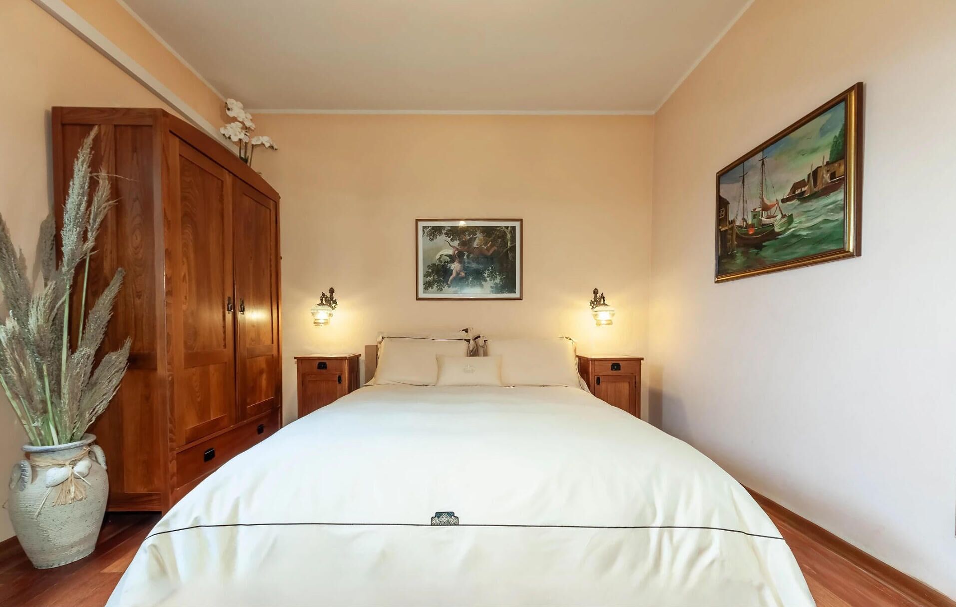 A bedroom with a large bed, wooden wardrobe, two bedside tables with lamps, a painting on the wall, and a potted plant.