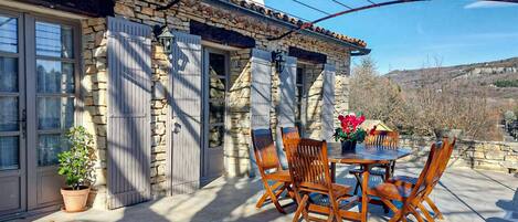 A stone building with a patio area featuring a wooden table and chairs, a potted plant, and a view of the surrounding landscape.