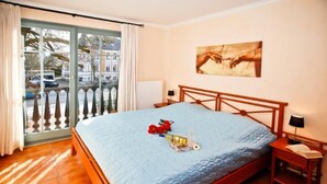 A bedroom with a large bed, a window with a view of buildings, a painting on the wall, and a balcony with a railing. - (293) 3-room apartment beach road - Apartment block LAGUNA (Kühlungsborn)