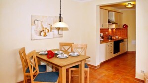 A dining area with a wooden table and chairs, a painting on the wall, and a kitchen in the background. - (293) 3-room apartment beach road - Apartment block LAGUNA (Kühlungsborn)