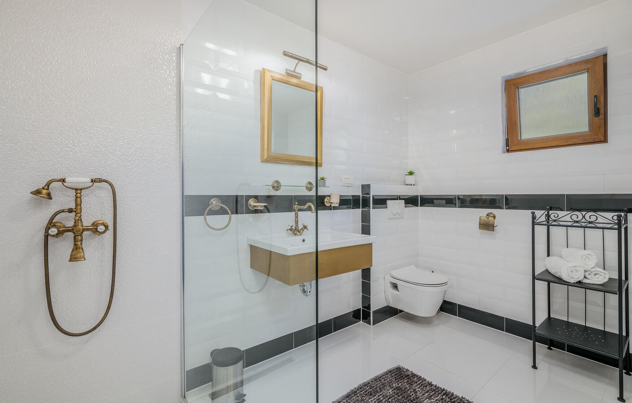 Ein modernes Badezimmer mit Duschkabine aus Glas, einem weißen Waschbecken, einer Toilette und einem schwarzen Metallregal mit Handtüchern.