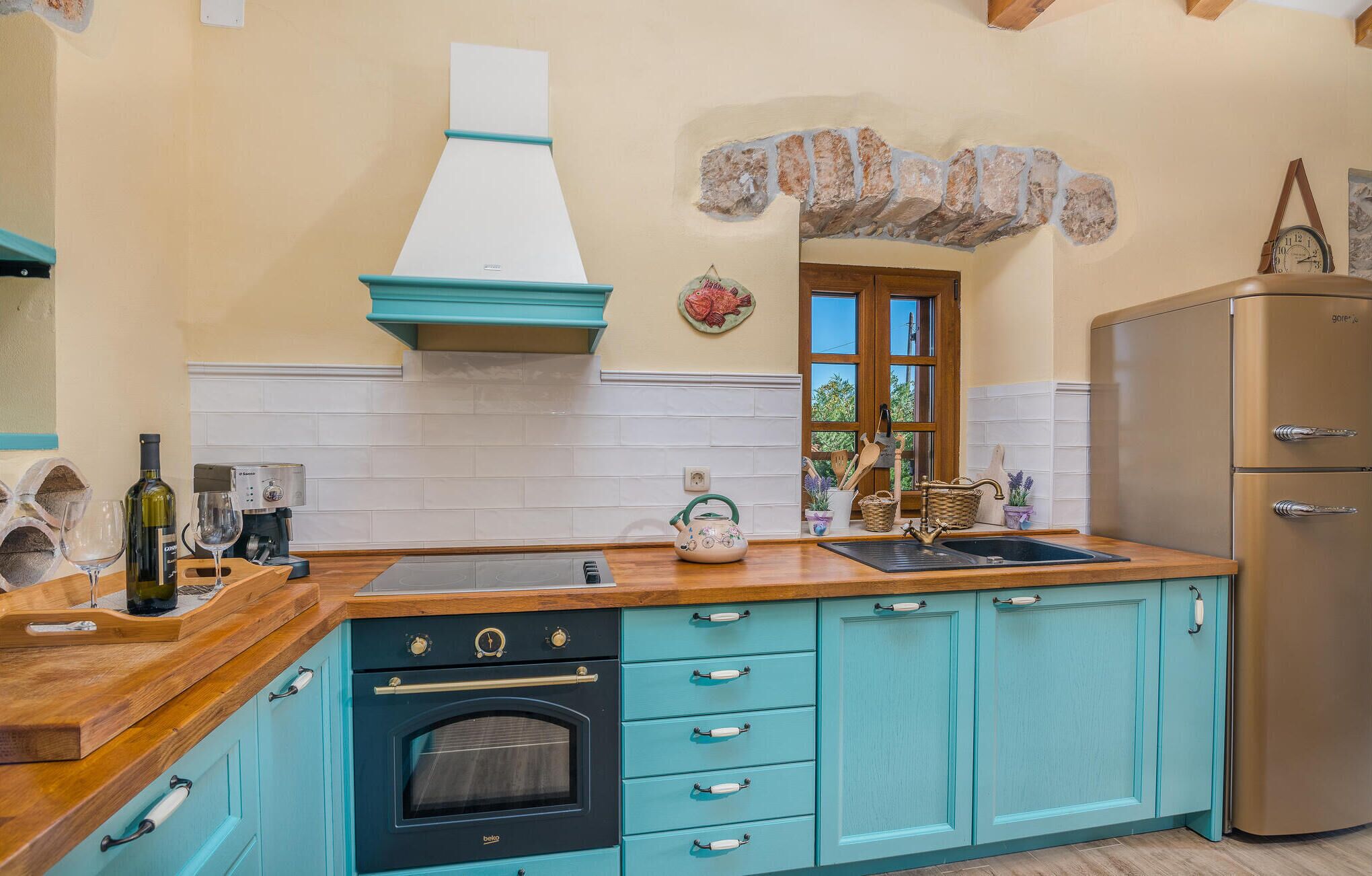 A kitchen with teal cabinets, a wooden countertop, a built-in oven, a stainless steel refrigerator, and a window with a view of greenery outside.