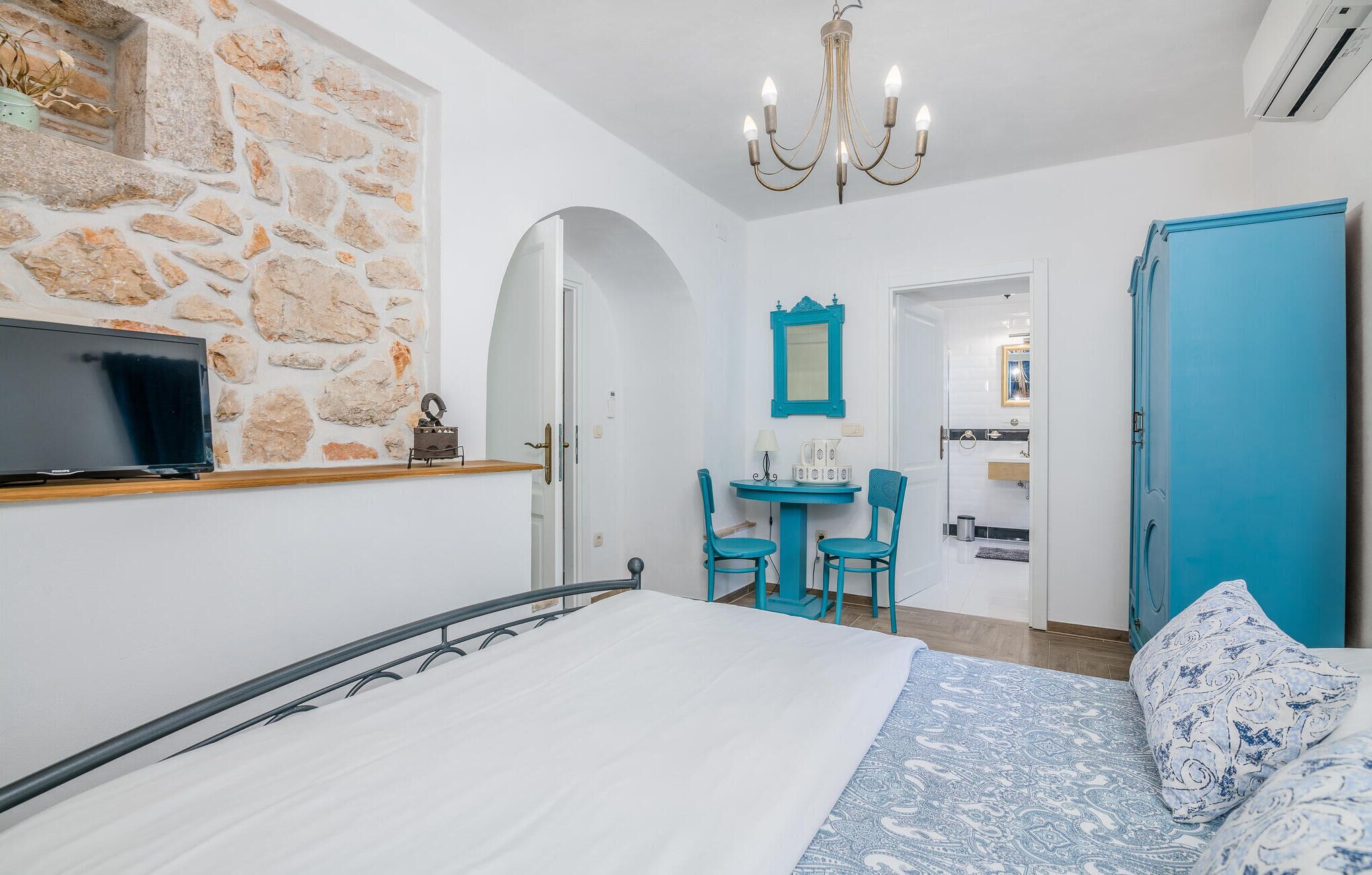 Une chambre avec un mur en pierre, un lit avec une literie blanche, une télévision, un lustre, une salle de bain visible à travers une arche, et une porte bleue.