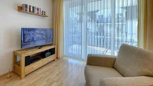 Uma sala de estar com uma TV sobre um suporte de madeira, um sofá, uma estante e uma janela grande com cortinas translúcidas.