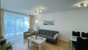 Uma sala de estar moderna com um sofá cinzento, uma mesa de centro branca e uma área de jantar com mesa de vidro e cadeiras pretas.