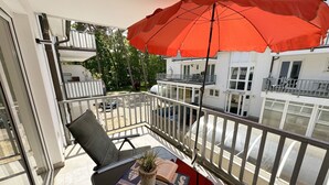 Uma varanda com um guarda-sol vermelho, uma mesa com uma planta e um livro, e vista para edifícios vizinhos.