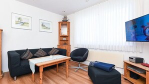 A living room with a black sofa, a wooden coffee table, a black armchair, a small ottoman, a TV, and a bookshelf. - Vacation apartment Treasure Island (Esens)