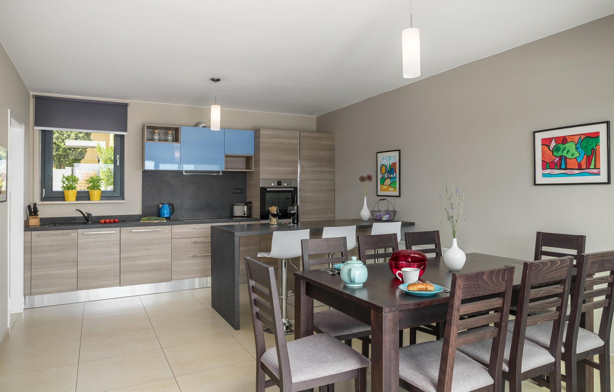 A modern kitchen with a central island, a dining area with wooden chairs, and a large window with potted plants.