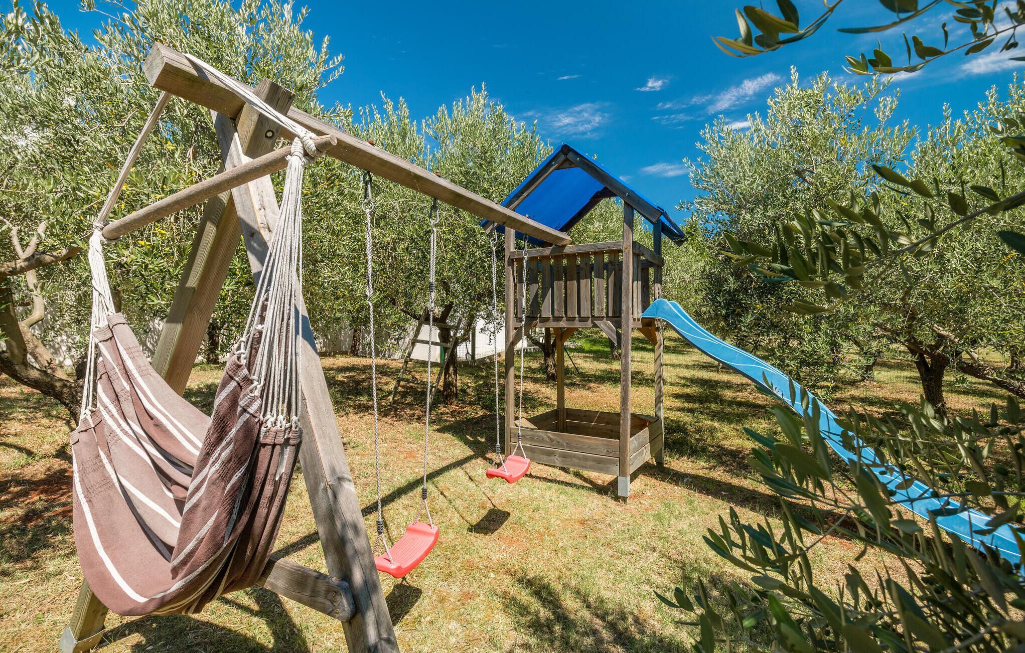 Un’altalena in legno con un’amaca, uno scivolo e una casetta da gioco in un’area erbosa con ulivi.