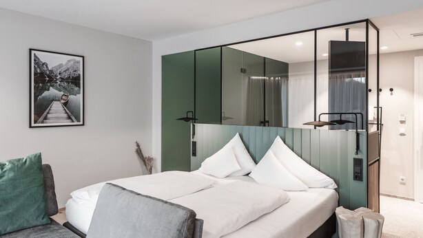 A modern hotel room with a large bed, a seating area with a sofa and armchair, a large mirror, and a framed picture on the wall. - Double Deluxe Plus - Hotel Goldenes Schiff (Bad Ischl)
