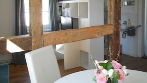 Ein Zimmer mit Holzbalken, weißen Wänden, einem weißen Stuhl, einem runden Tisch mit Blumenvase, einem Bücherregal und einem Badezimmer mit Waschbecken und Spiegel.