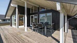 A wooden deck with a table and chairs, a covered patio area, and large windows. - Beach front home in Ringkøbing with sauna (Ringkøbing)