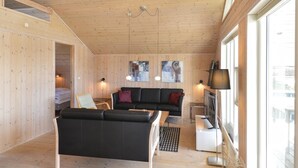 A wooden interior with a black sofa, a small table, and a TV. There are two wall-mounted lights and a ceiling fan. - Beach front home in Ringkøbing with sauna (Ringkøbing)