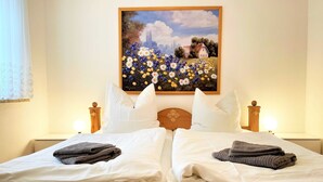A double bed with white linens and two pillows, a painting of a field with flowers and a house on the wall, and bedside lamps. - Tom and Jerry Vacation Rental - Silberseehaus in the Mortka Recreation Oasis (Lohsa)