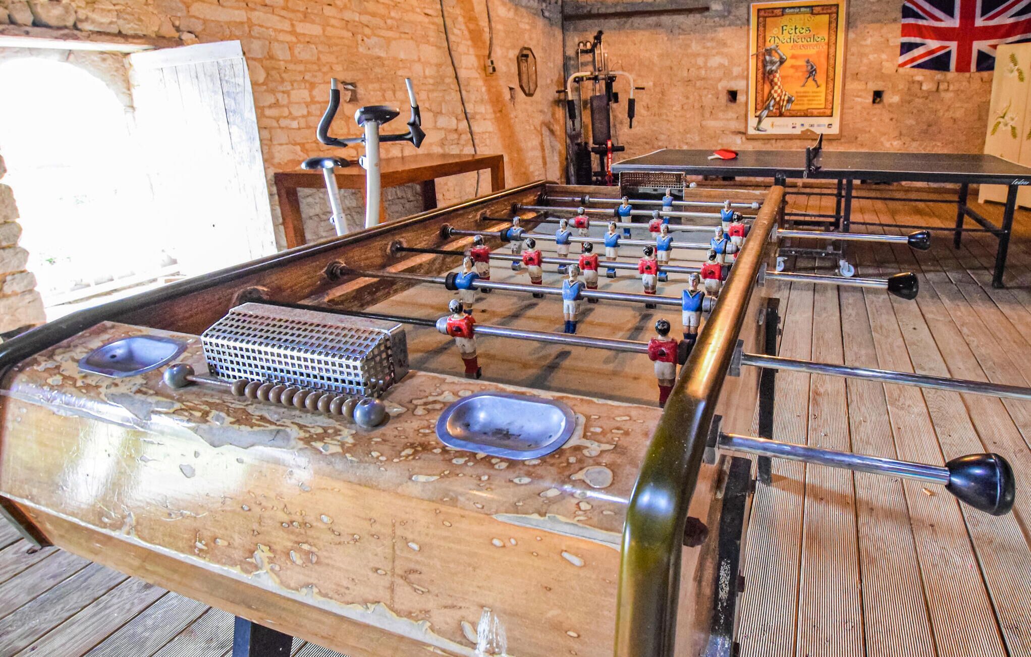 A foosball table in a room with brick walls, a wooden floor, and a table tennis table in the background.