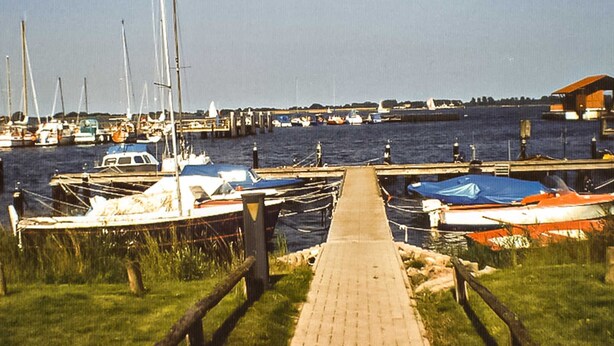 A marina with boats docked, a wooden walkway leading to the water, and a distant pier. - Holiday Bungalow - Bungalow (Reinberg)