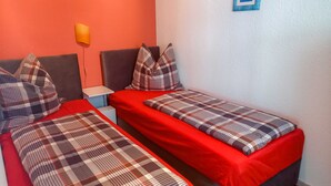 A room with two beds, each with red bedding and plaid-patterned pillows. There is a small bedside table with a lamp on the left side. - Holiday Bungalow - Bungalow (Reinberg)