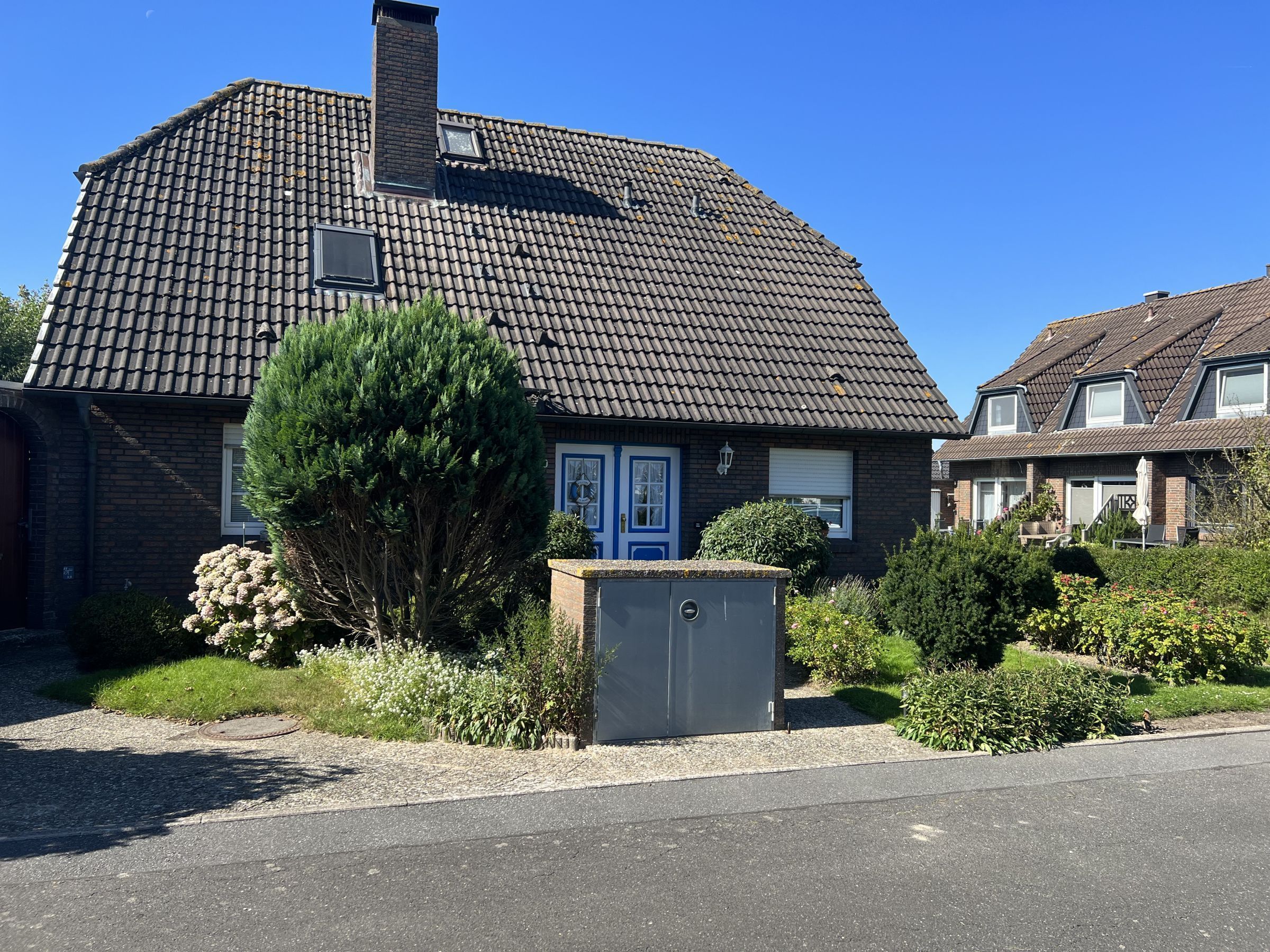A two-story house with a tiled roof, a blue door, and a small garden with a tree and shrubs.
