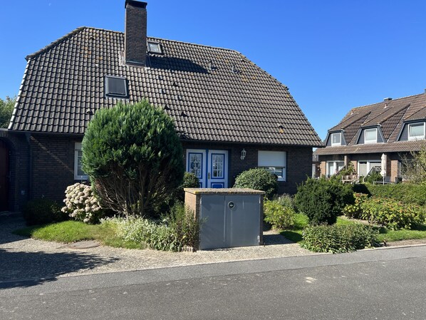 A two-story house with a tiled roof, a blue door, and a small garden with a tree and shrubs.